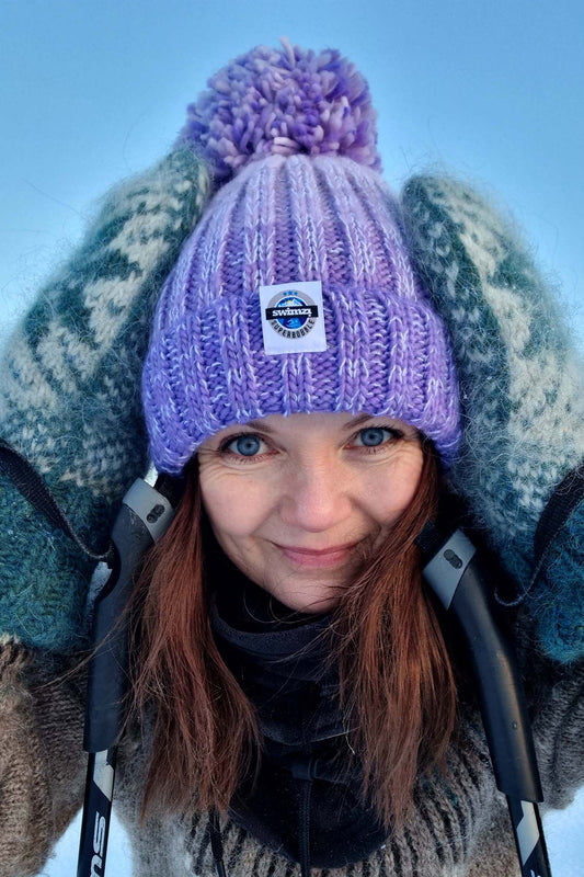 Woman standing, with her hands to her head, against the bright blue sky of Norway wearing a chunky knit Super Bobble hat in shades of lavender. The hat has a big pom-pom and a deep ribbed headband. It contains reflective yarns and is lined in luxury sherpa fleece.