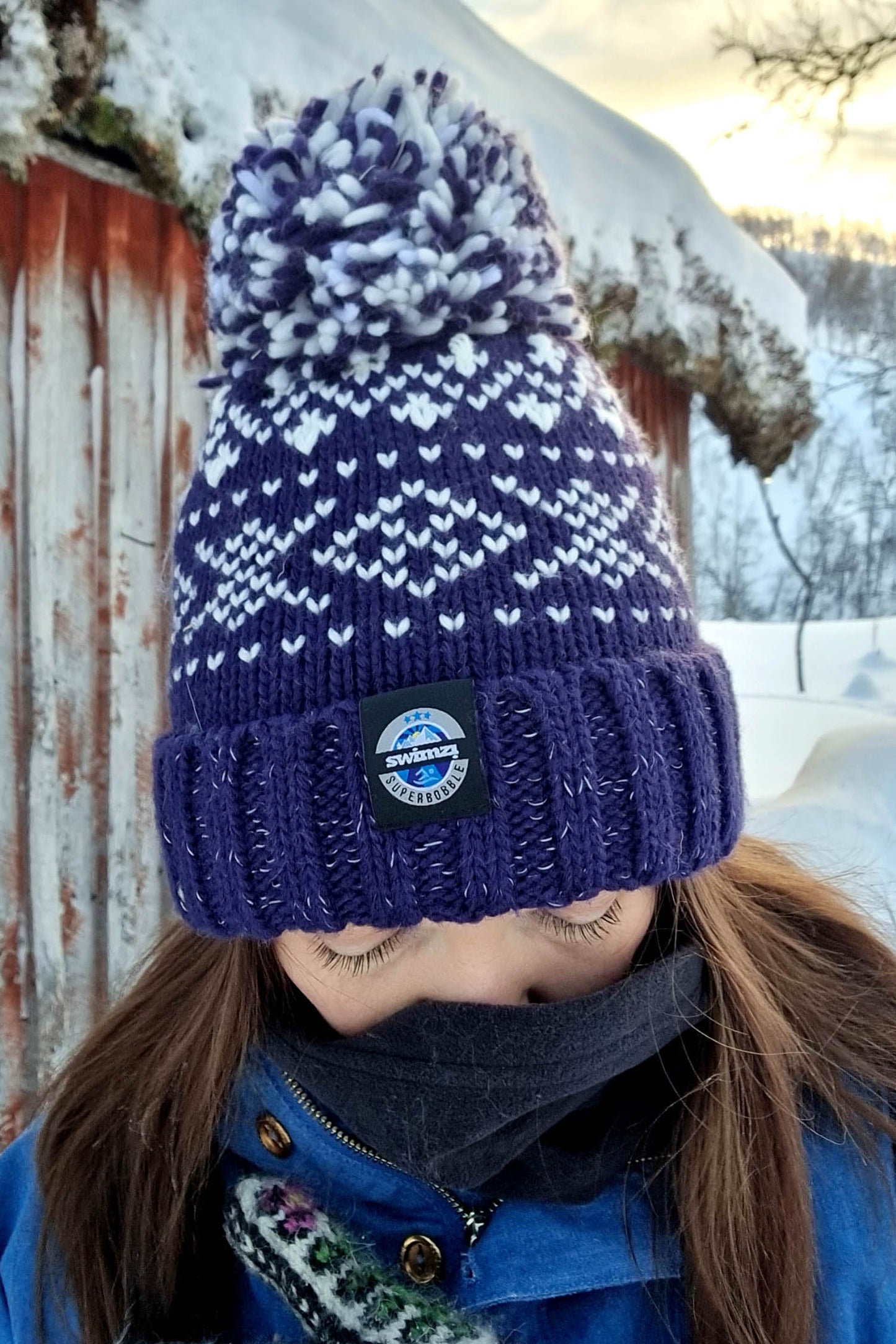 Woman at the side of a log cabin in Norway is wearing a chunky Nordic knit bobble hat. The navy and white Nordic design hat also has a deep ribbed headband, contains reflective yarns and has a luxury sherpa fleece lining.
