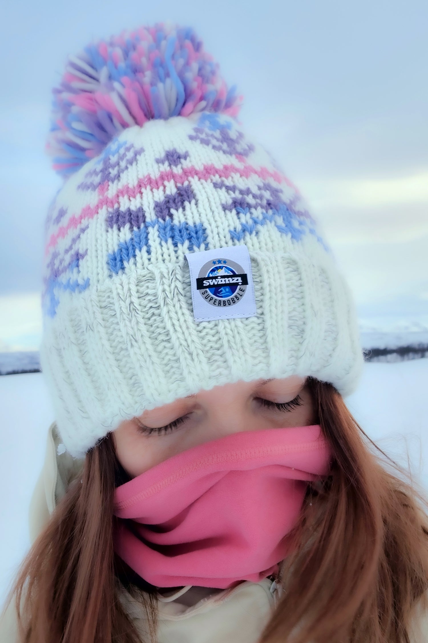 Woman in the snowy foothills of Northern Norway is wearing a chunky Nordic knit bobble hat. The white base and cool pastel toned Nordic design hat also has a deep ribbed headband, contains reflective yarns and has a luxury sherpa fleece lining.