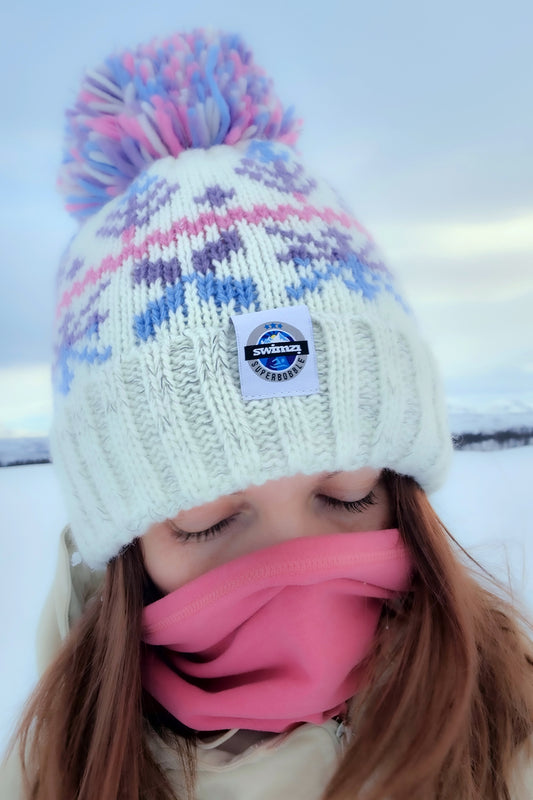 Woman in the snowy foothills of Northern Norway is wearing a chunky Nordic knit bobble hat. The white base and cool pastel toned Nordic design hat also has a deep ribbed headband, contains reflective yarns and has a luxury sherpa fleece lining.