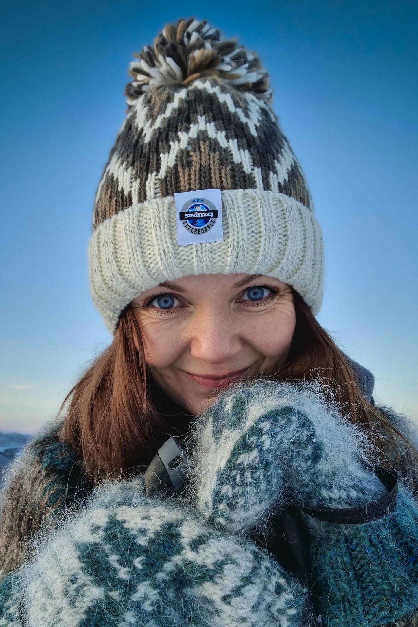 Set against a beautiful blue sky in Norway woman is wearing a Super Bobble hat in cream and soft brown shades in a zig-zag design, The hat  has a deep ribbed headband, contains reflective yarns and has a luxurious sherpa fleece lining.