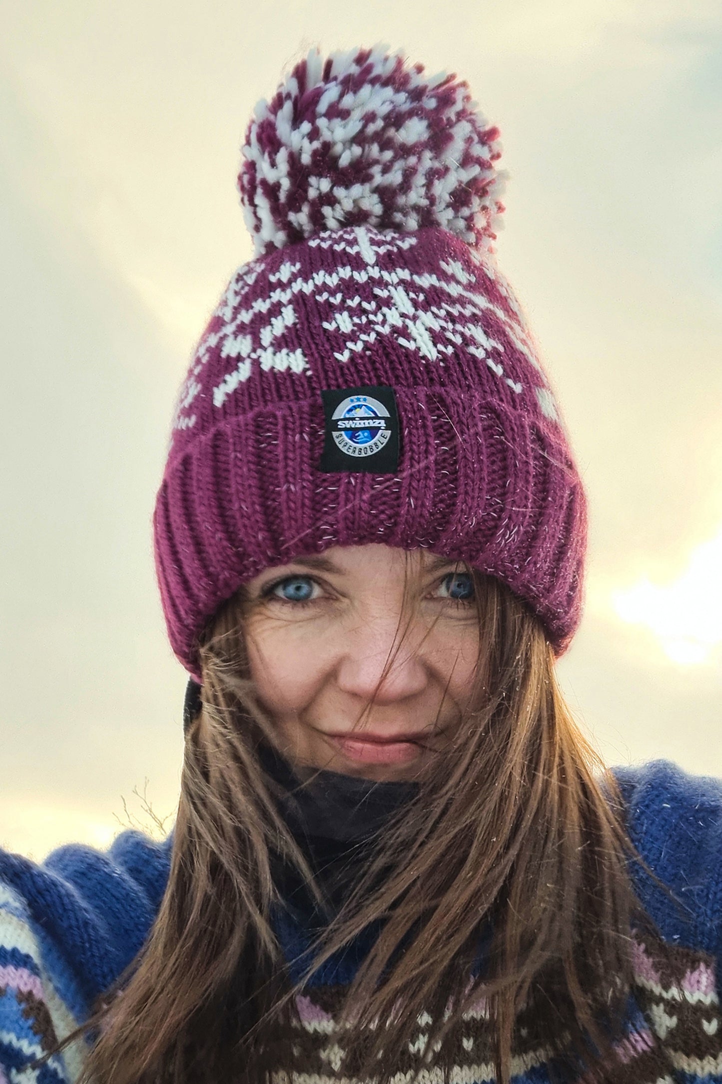 Woman sitting on the snow of Northern Norway is wearing a chunky Nordic knit bobble hat. The burgundy red base and white Nordic design hat also has a deep ribbed headband, contains reflective yarns and has a luxury sherpa fleece lining.