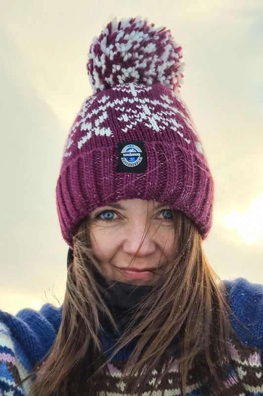 Woman sitting on the snow of Northern Norway is wearing a chunky Nordic knit bobble hat. The burgundy red base and white Nordic design hat also has a deep ribbed headband, contains reflective yarns and has a luxury sherpa fleece lining.