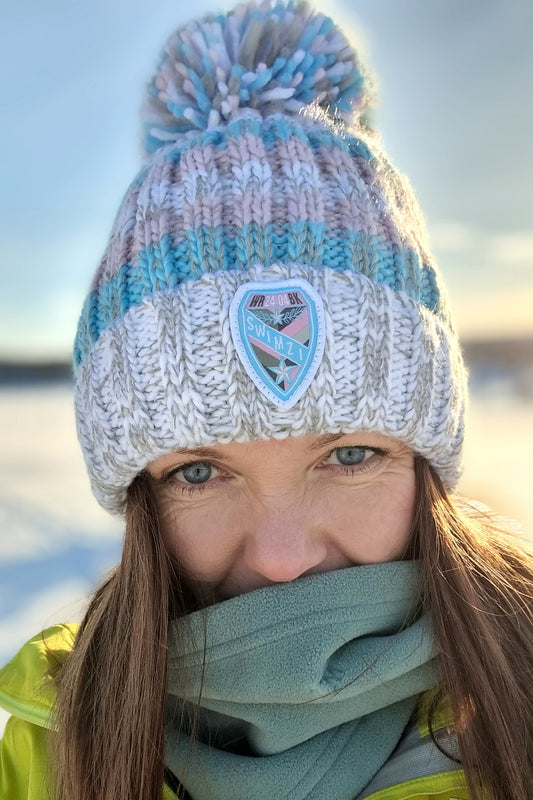 Set against a snowy arctic landscape woman is wearing a white and light grey based super bobble hat with cool pastel pink and blue stripes.  The hat has a deep ribbed headband, contains reflective yarns and is lined in a luxurious sherpa fleece.