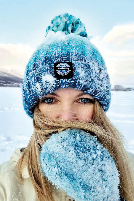 Woman stands amongst a snowy landscape, wearing a striped bobble hat in a  shades of dark to light turquoise blue with a large pom-pom. The hat also has a deep ribbed headband and contains reflective yarns and a luxury sherpa fleece lining.