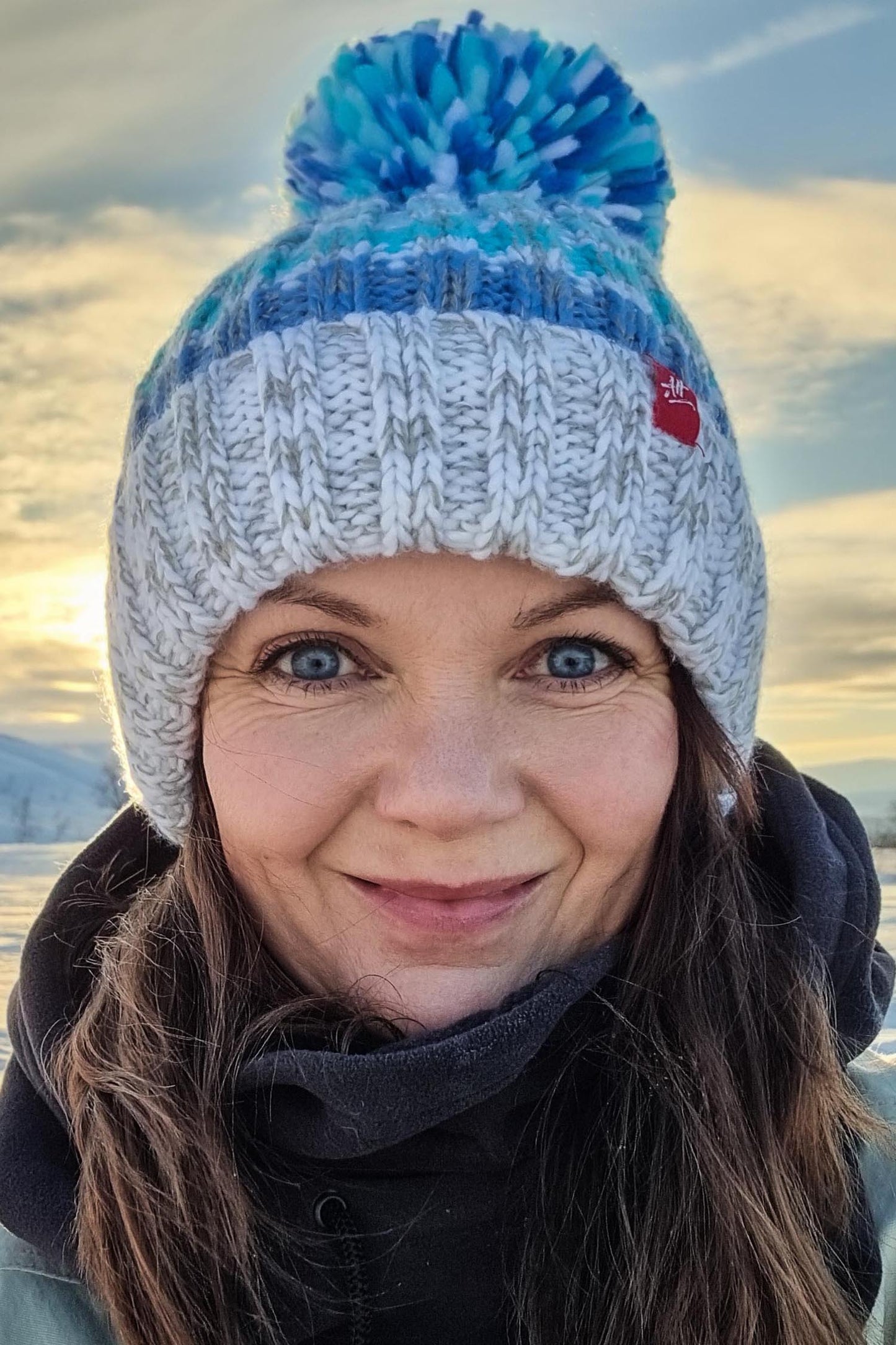  Woman against the backdrop of snow is wearing a Super Bobble hat with a base of white and pale grey and shades of sea blue stripes. The hat has a deep ribbed headband, contains reflective yarns andis lines in luxurious sherpa fleece.