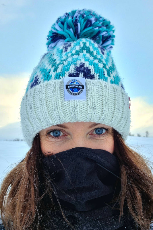 Woman stands amongst the crisp white arctic landscape, wearing a bobble hat in a zig-zag design in white and shades of blue with large pom-pom. The hat also has a deep ribbed headband and contains reflective yarns and a luxury sherpa fleece lining.