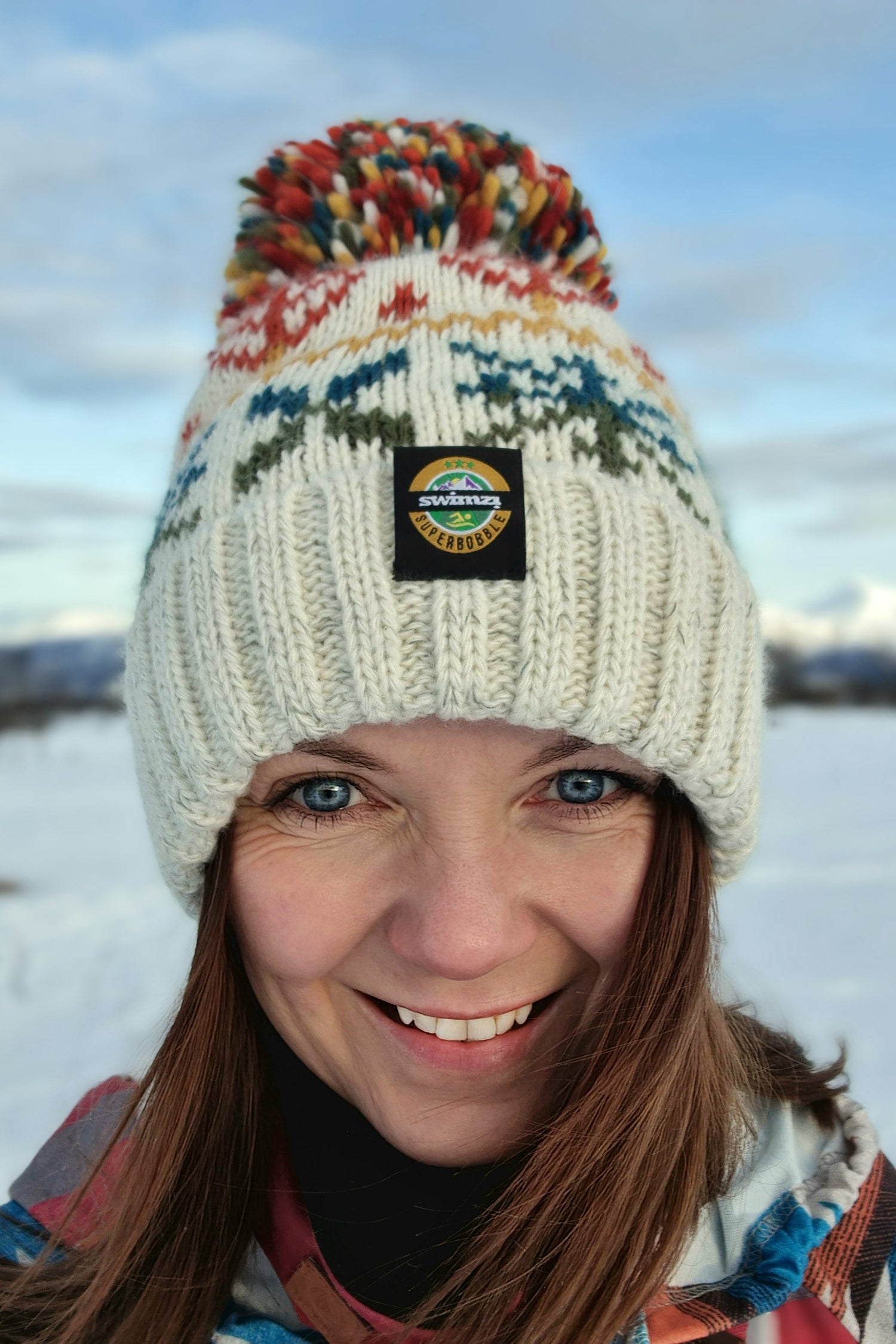 Woman in the snowy foothills of Northern Norway is wearing a chunky Nordic knit bobble hat. The oatmeal base and warm autumn toned Nordic design hat also has a deep ribbed headband, contains reflective yarns and has a luxury sherpa fleece lining.