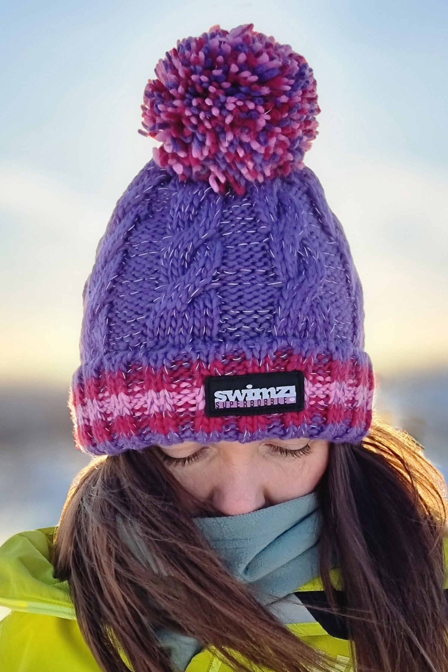 Women wearing a purple cable knit super boble hat with a deep ribbed headband which is striped in deep and light pink shades agains t the purple reminiscent of berry shades. The hat is also has a reflective thread running through and is lined with a luxurious sherpa fleece.