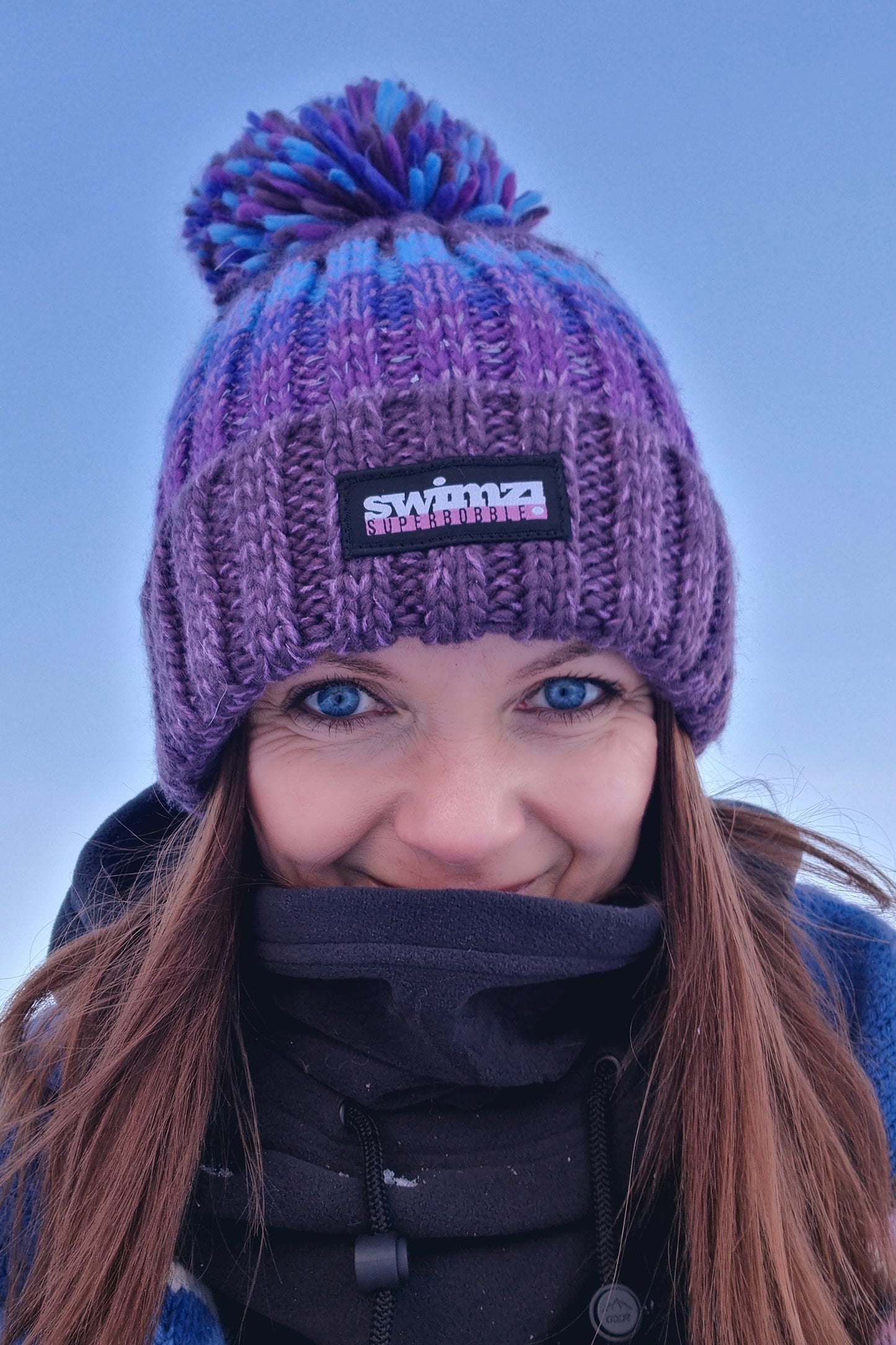  Woman standing in a snowy landscape in Norway is wearing a chunky knit Super Bobble hat in berry shades of purple and blue. The hat has a big pom-pom and a deep ribbed headband. It contains reflective yarns and is lined in luxury sherpa fleece. 