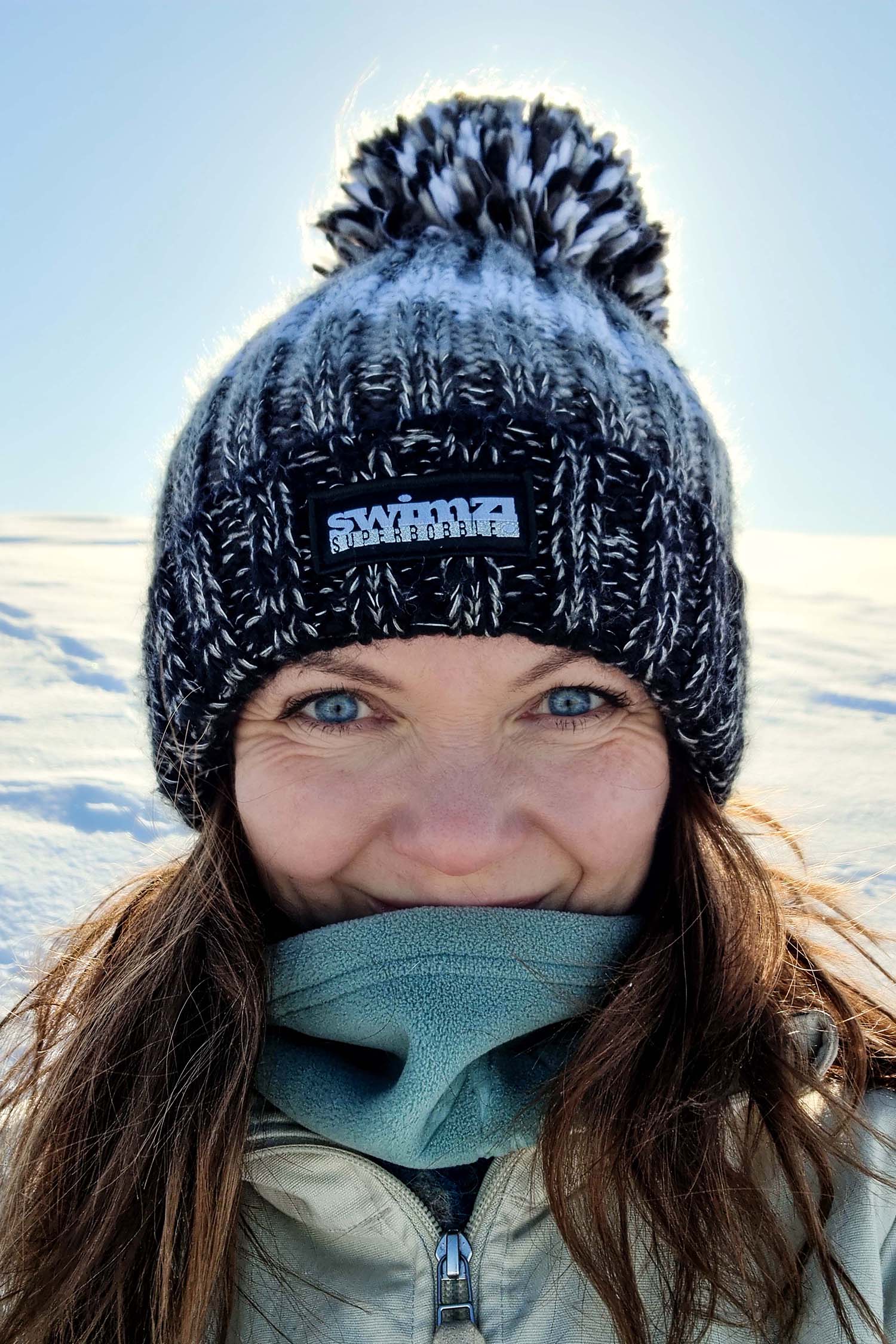 Woman walking through the snow-covered hills of Norway in a chunky knit Super Bobble hat in black, charcoal and white. The hat has a big pom-pom and a deep ribbed headband. It contains reflective yarns and is lined in luxury sherpa fleece.