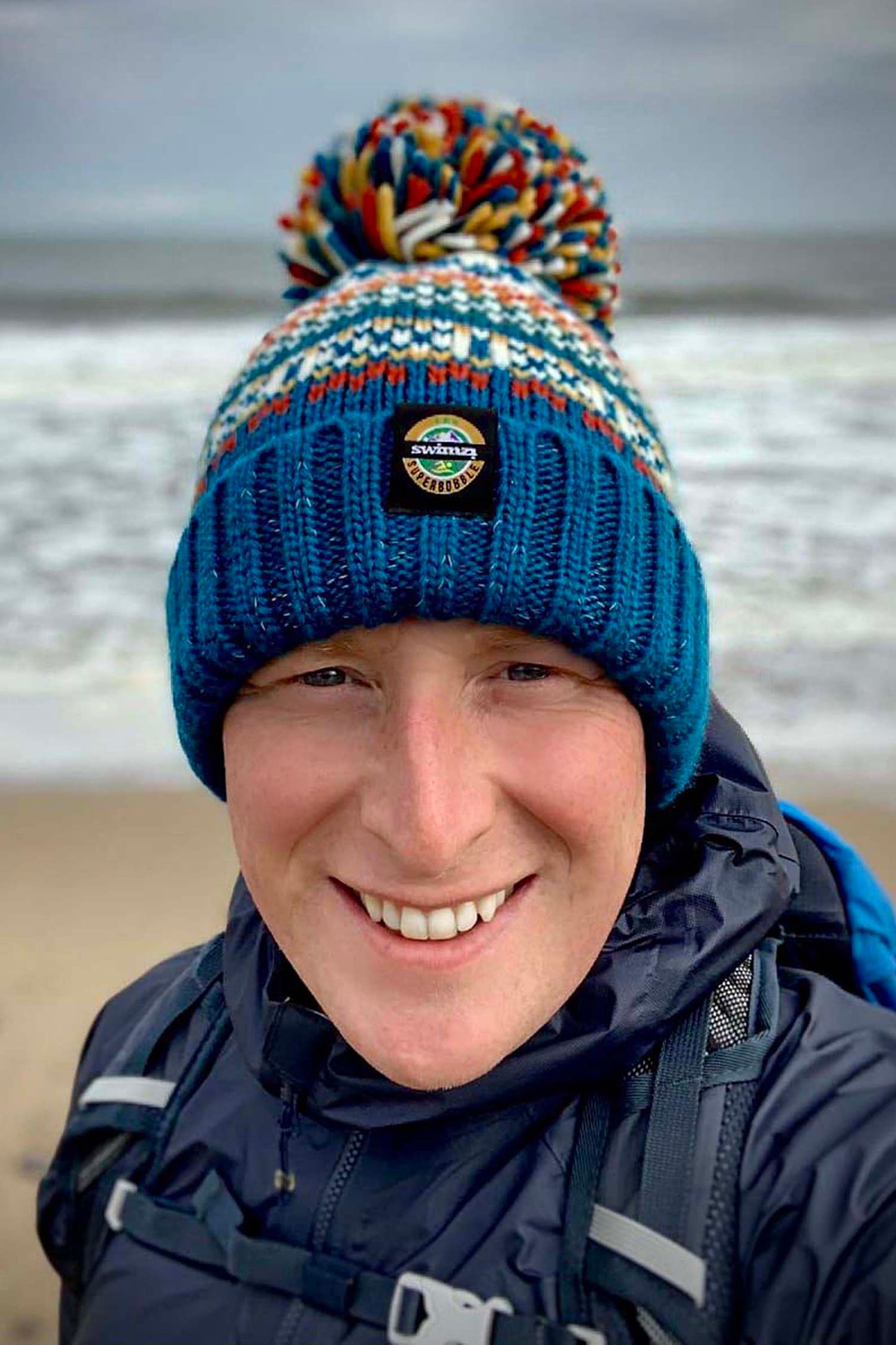 Man standing on the rugged coastline is wearing a chunky knit Nordic style Super Bobble hat in fjord blue, deep ochre and cream. The hat has a big pom-pom and a deep ribbed headband, contains reflective yarns and is lined in luxury sherpa fleece.