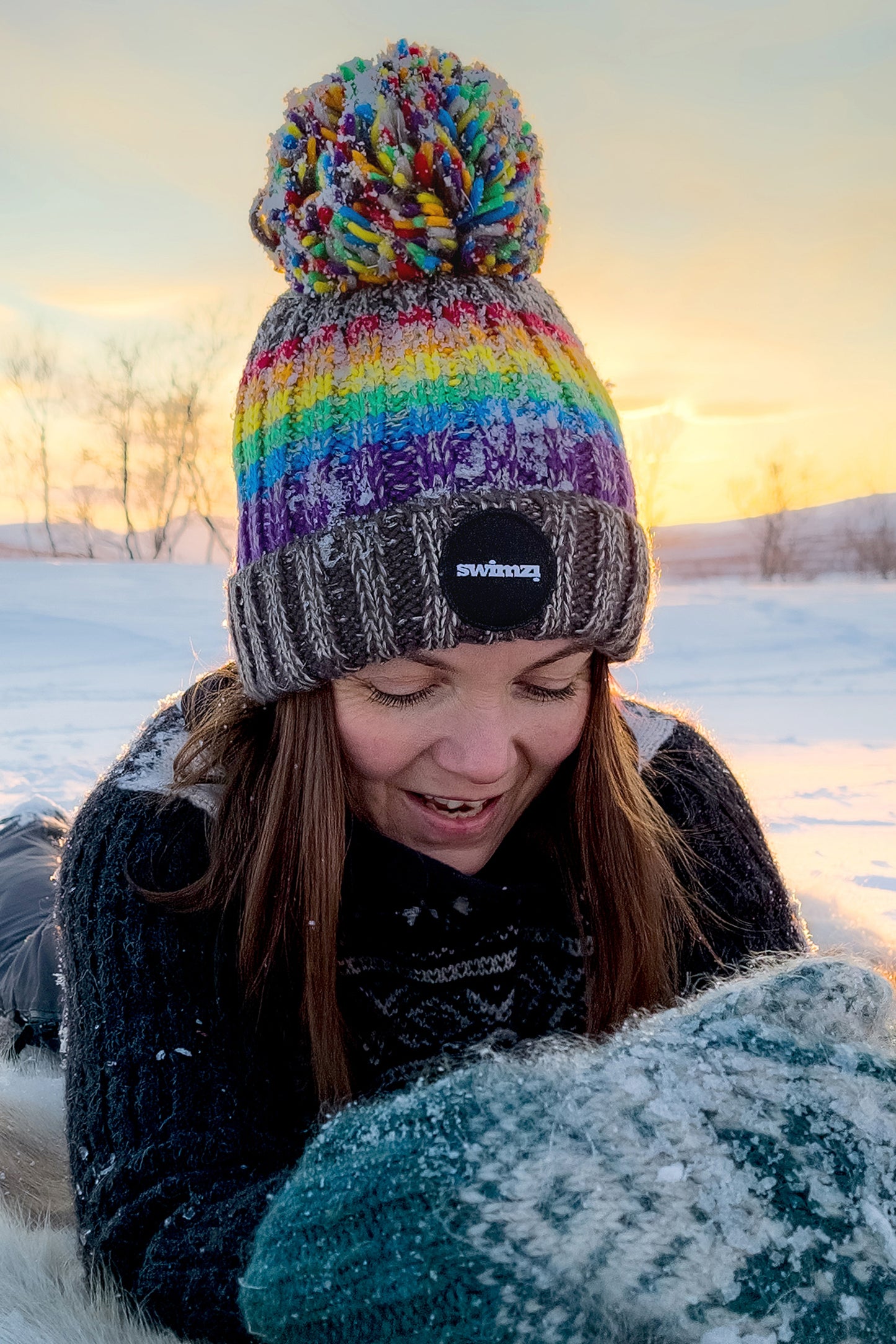 Woman laying on the Norwegian snow is wearing a chunky rib knit bobble hat in rainbow coloured stripes. The hat also has a deep ribbed headband, contains reflective yarns and has a luxury sherpa fleece lining.