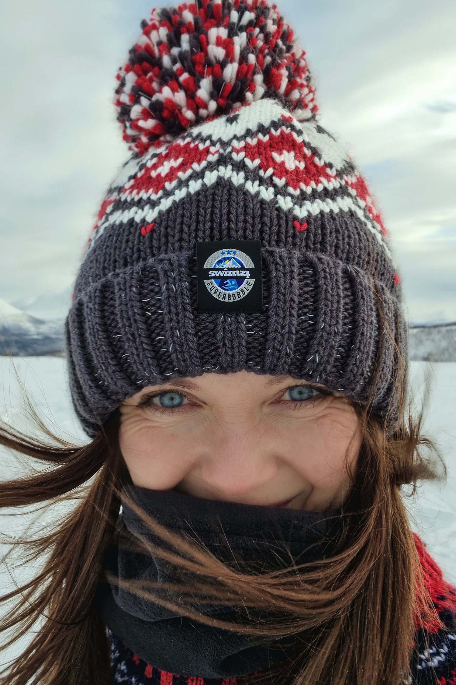 The snow of winter provide the backdrop for woman who is wearing a chunky Nordic knit Super Bobble hat in dark grey with white and red Tyrol design. The hat also has a deep ribbed headband, contains reflective yarns and has a luxury sherpa fleece lining.