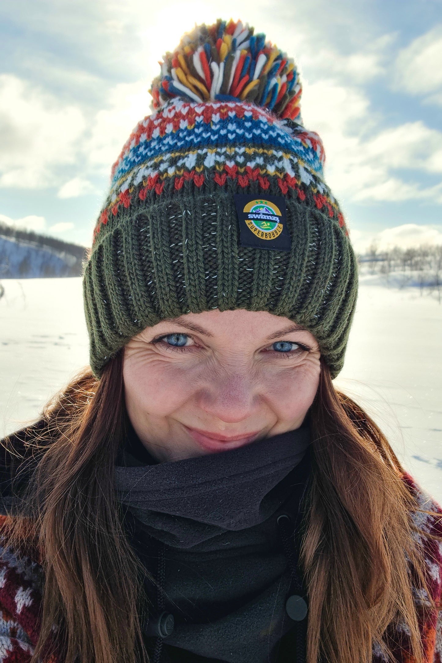 Woman against the icy landscape is wearing a  chunky  knit bobble hat in a dark olive green shade with a nordic design in shades of  white, blue, burnt orrange and mustard yellow. The hat has a big pom-pom and a deep ribbed headband, contains reflective yarns and is lined in luxury sherpa fleece.