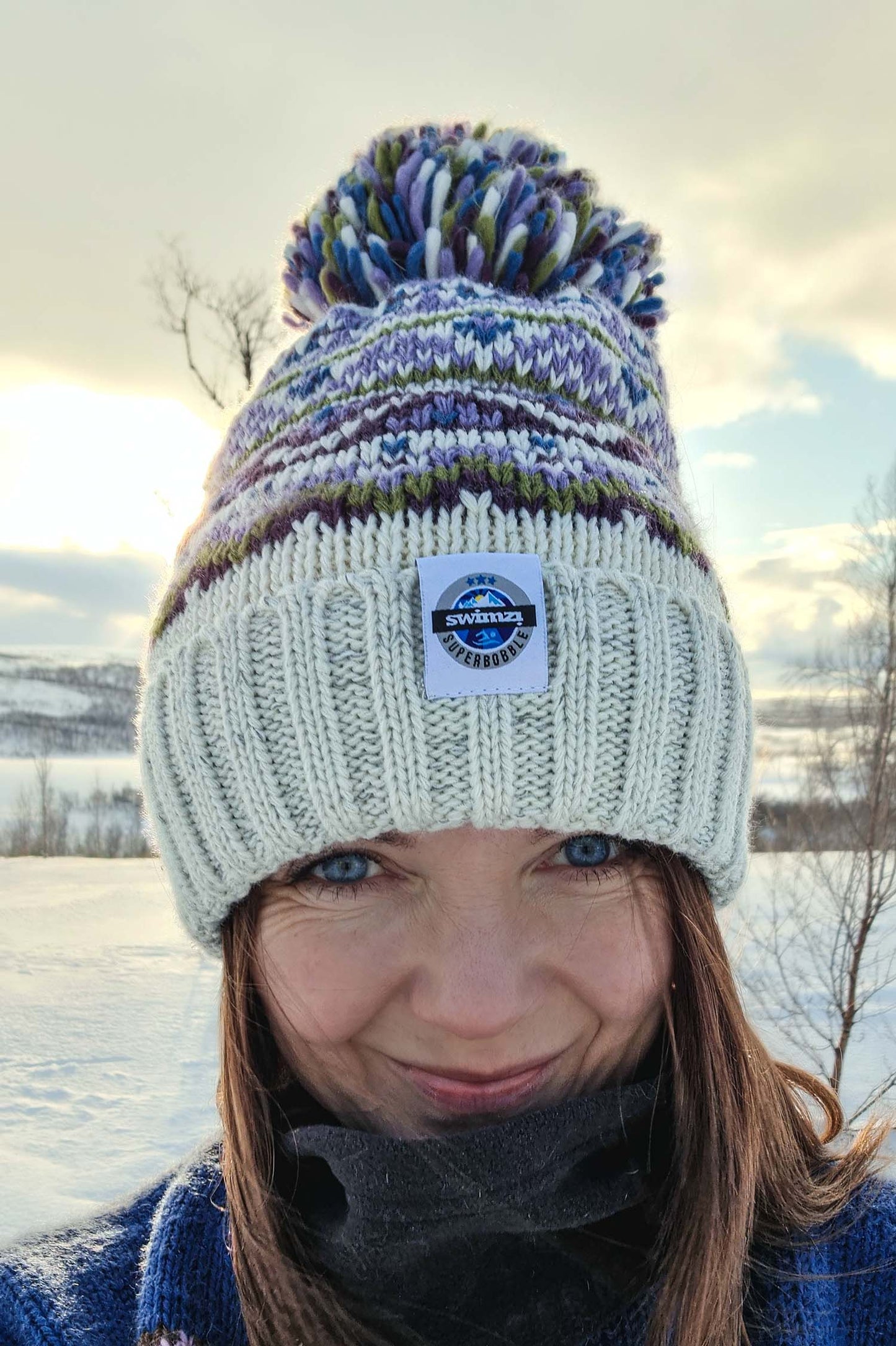 Woman in the Norwegian sun fading over the horizon is wearing a white based bobble hat with heather and moss green traditional fairisle pattern.  The hat also has a deep ribbed headband and contains reflective yarns and a luxury sherpa fleece lining.