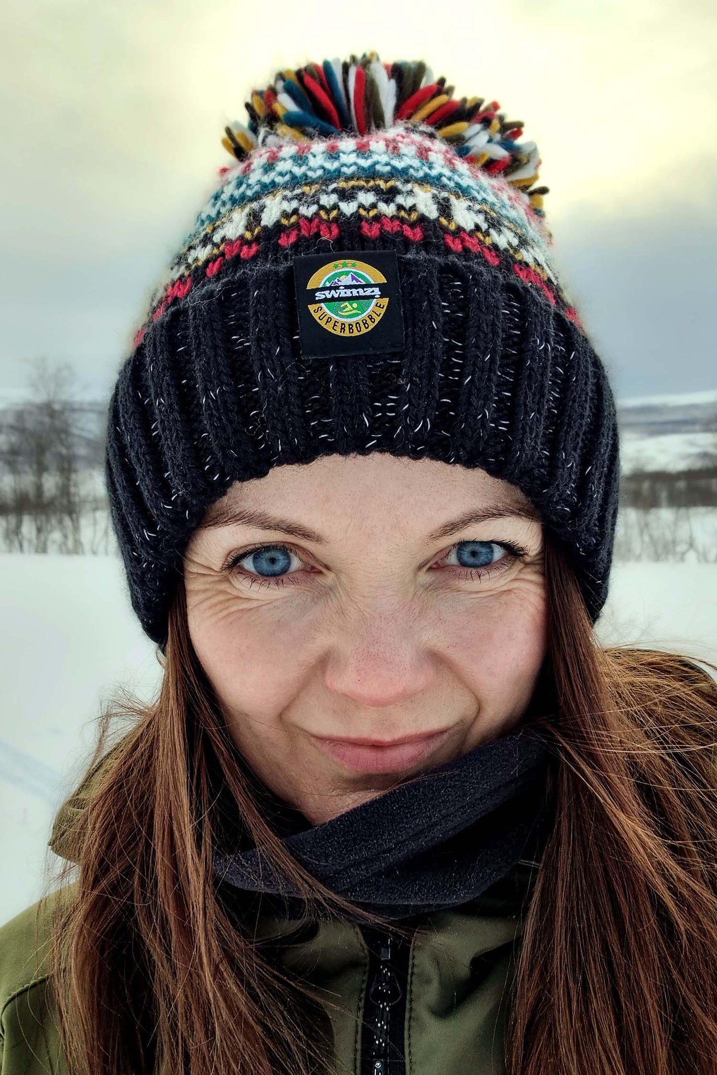 Woman in a wintery landscape backlit by a fading sunset is wearing a traditional Nordic design chunky knit Super Bobble Hat. The black base knit is accented by dark red, deep teal and warm cream.