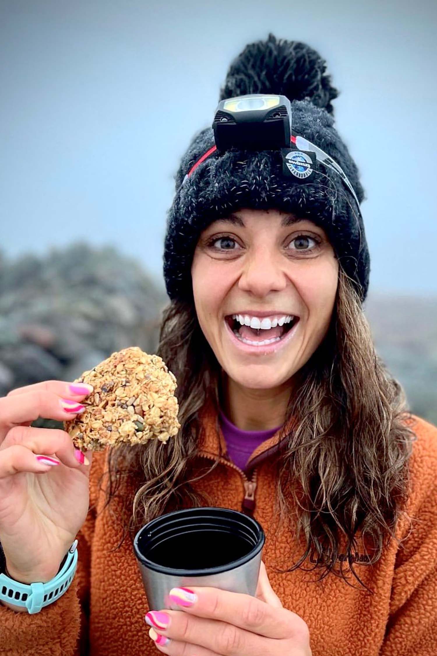 Woman at the summit of a mountain is wearing a chunky cable knit Super Bobble hat in jet black. The hat also has a deep ribbed headband and contains reflective yarns and a luxury sherpa fleece lining.