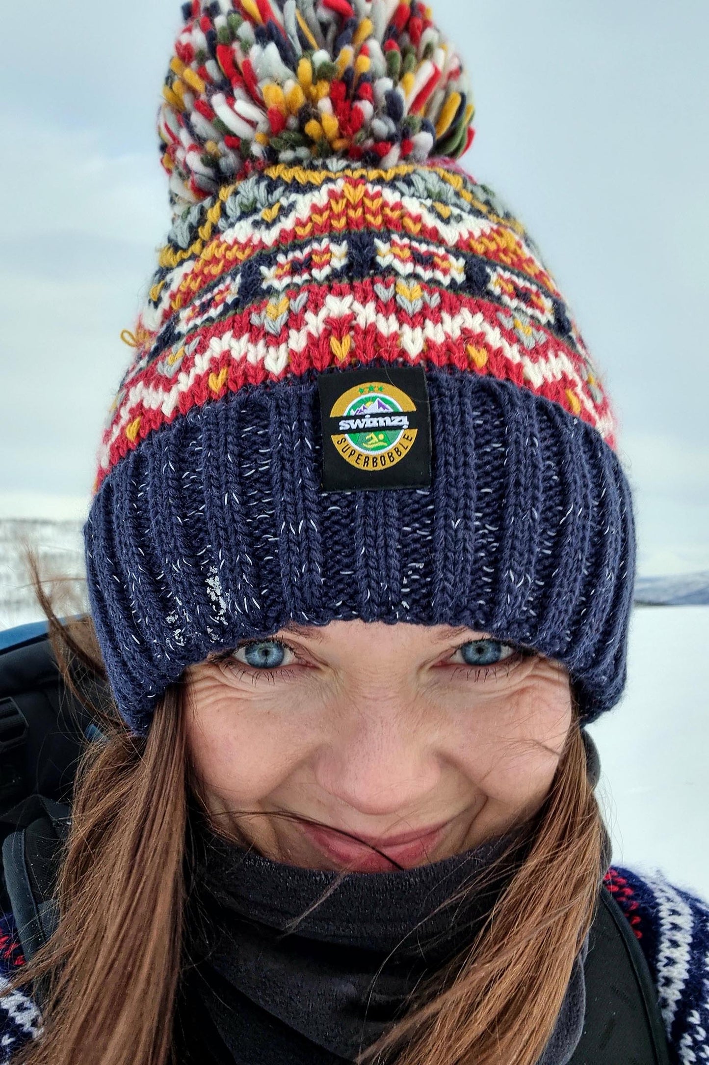 Woman in a snowy landscape is wearing a chunky fairisle knit Super bobble hat navy, red and mustard design. The hat also has a deep ribbed headband and contains reflective yarns and a luxury sherpa fleece lining.