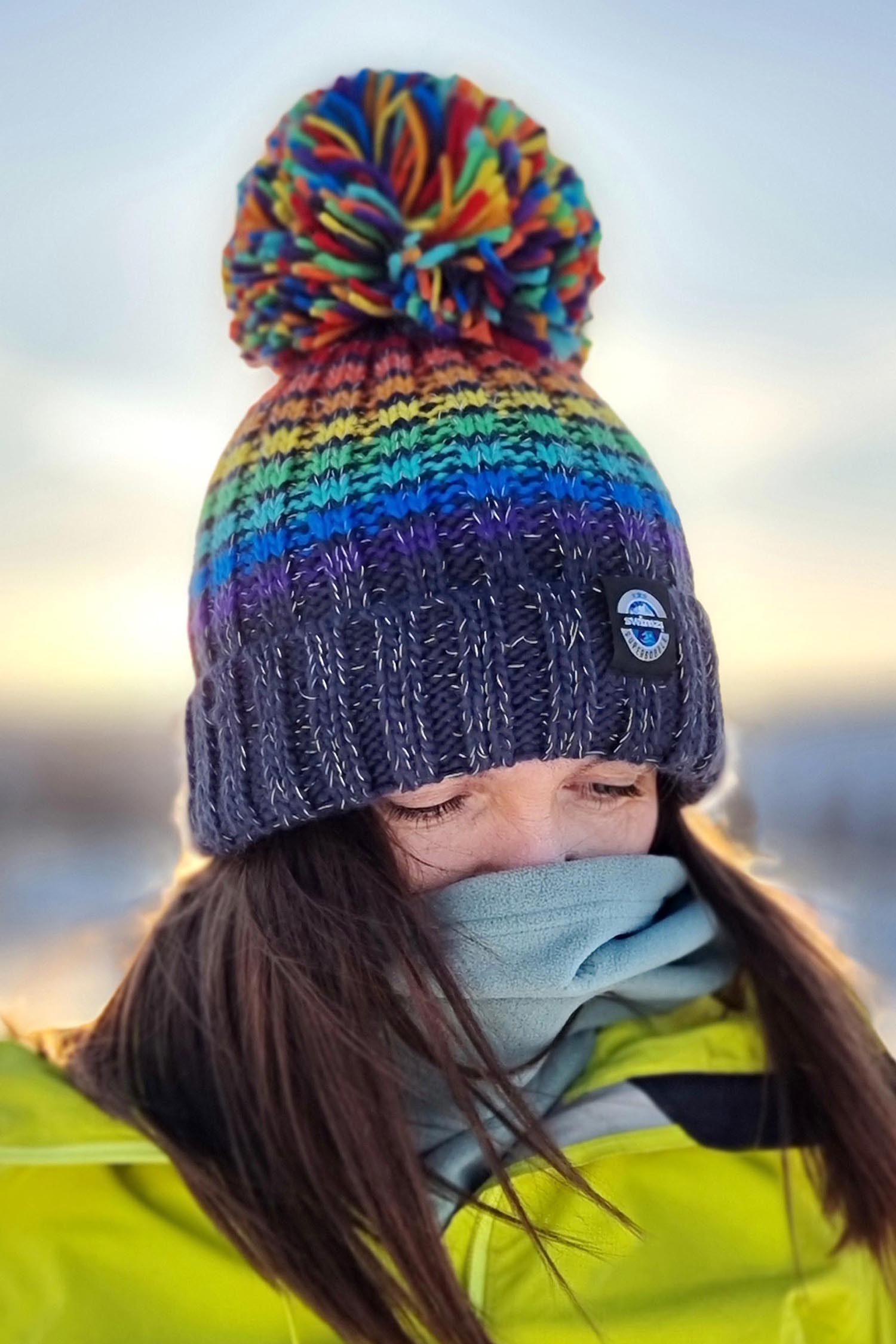 Woman wearing a chunky rib knit bobble hat in rainbow coloured stripes on a navy blue base, The hat also has a deep ribbed headband and contains reflective yarns with a luxury sherpa fleece lining.