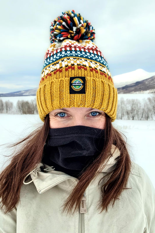 Woman in the icy winds punctuates the grey landscape with the bright mustard coloured chunky Nordic knit bobble hat she is wearing. The hat has a big pom-pom and a deep ribbed headband, contains reflective yarns and is lined in luxury sherpa fleece.