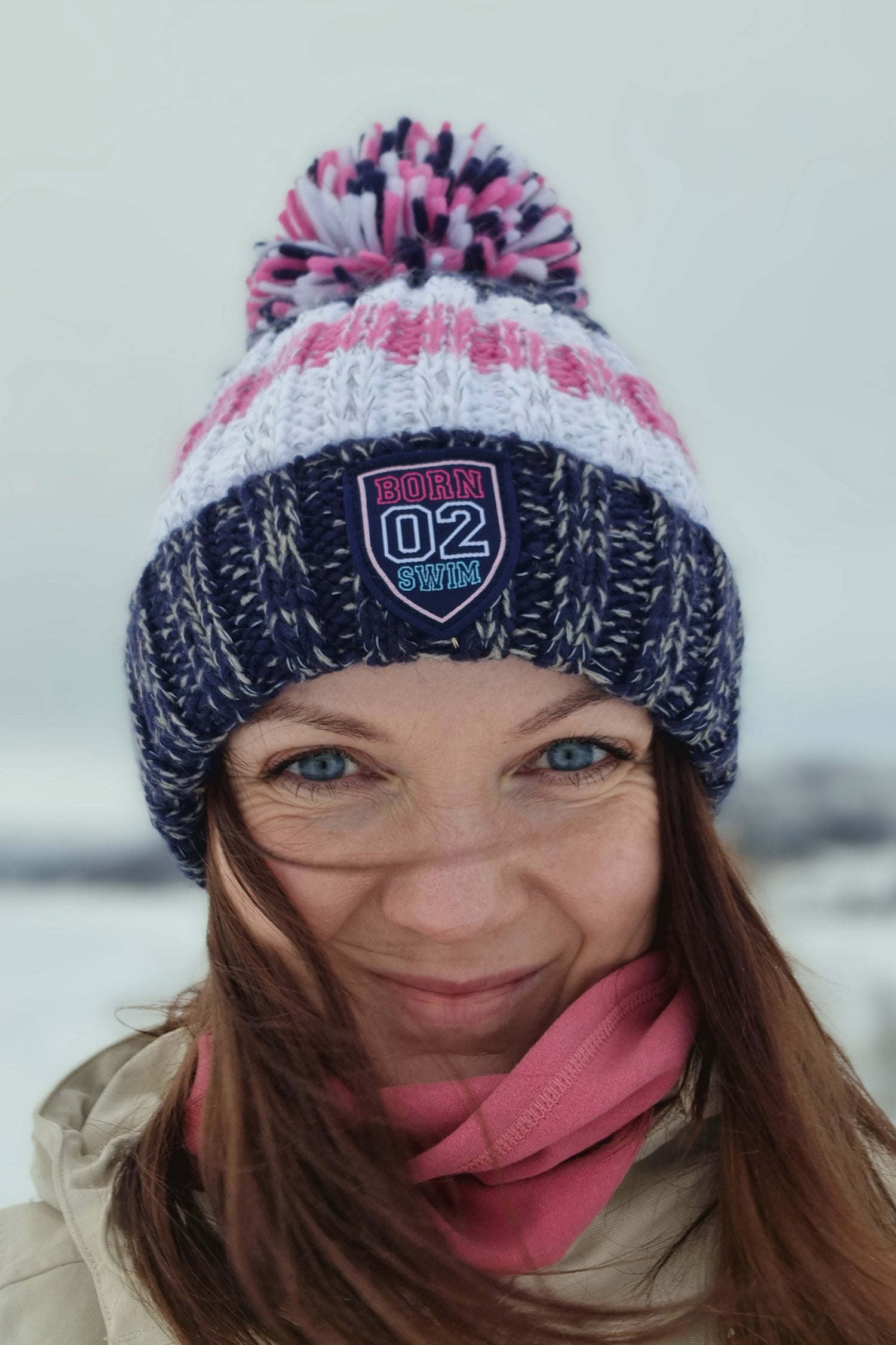 Woman in snowy tree lined glade in Northern Finland is wearing a navy, white and pale pink striped chunky knit Super Bobble hat.  The hat also has a deep ribbed headband and contains reflective yarns and a luxury sherpa fleece lining.