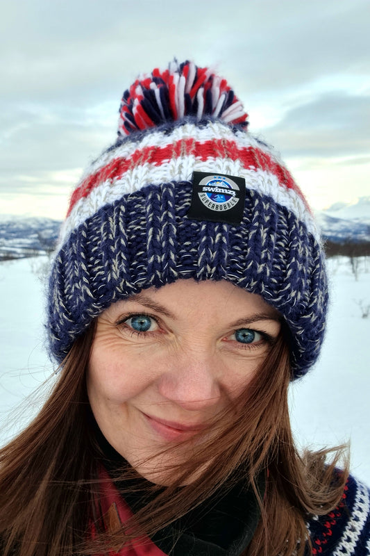 Women in a Scandinavian frozen landscape is wearing a stylish dark navy, white and red striped chunky Super Bobble hat with a large pom-pom and a deep ribbed headband. The hat contains reflective yarns and is lined in luxury sherpa fleece.