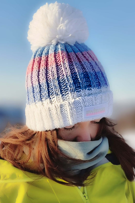  Woman standing in a snowy landscape in Norway, lit by the bright blue skies,  is wearing a chunky knit Super Bobble hat in graded shades of blue and pink. The hat has a big pom-pom and a deep ribbed headband. It contains reflective yarns and is lined in luxury sherpa fleece.