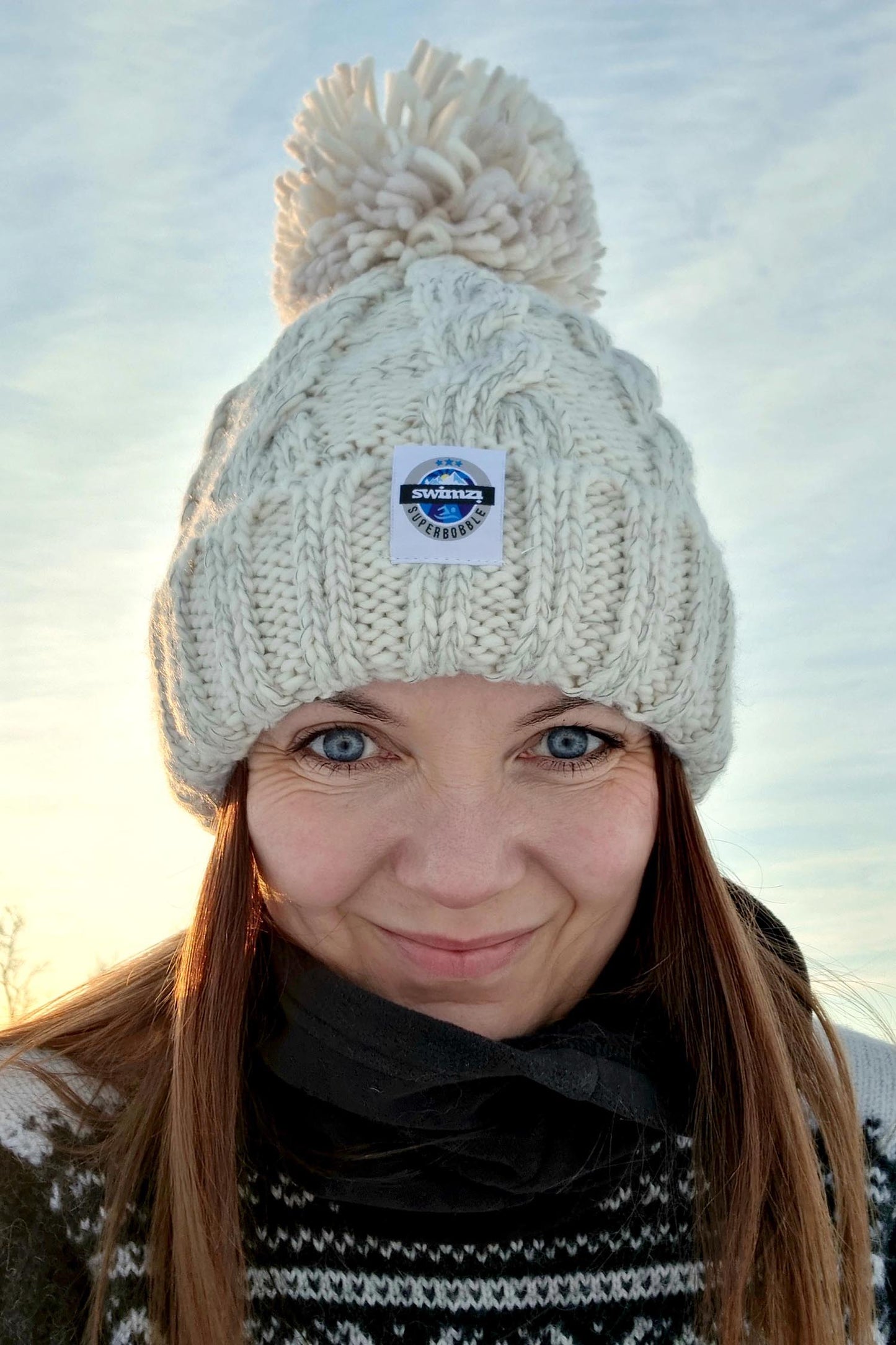 Woman in a snowy tree lined glade is wearing a cream oatmeal chunky cable knit Super Bobble hat. The hat also has a deep ribbed headband and contains reflective yarns and a luxury sherpa fleece lining.