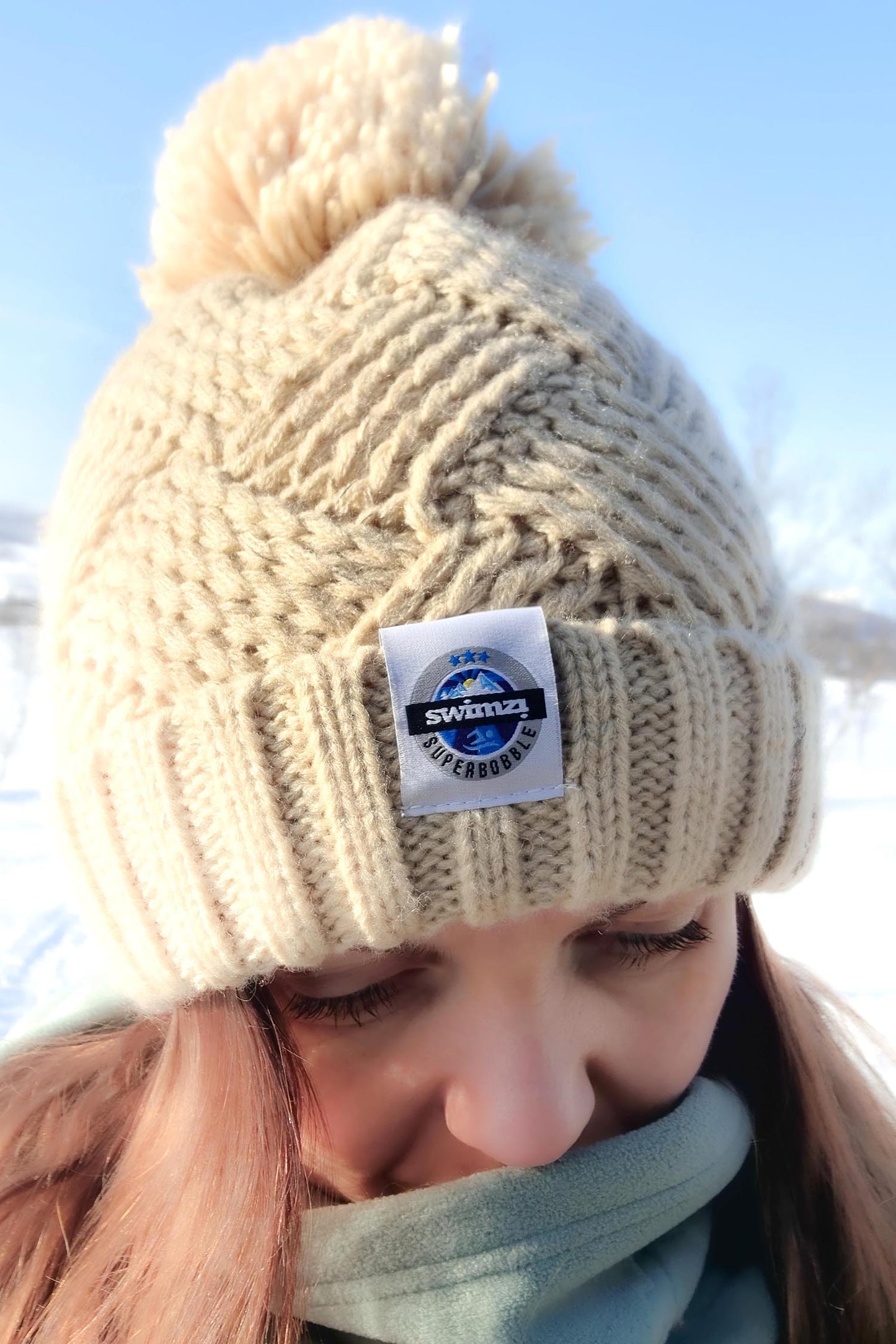 Woman cross country skiing through Norwegian wooded landscape wearing a chunky oatmeal bobble hat. It is knitted in an attractive cross patch pattern with a deep ribbed headband. The hat contains reflective yarns and is lined in luxury sherpa fleece.