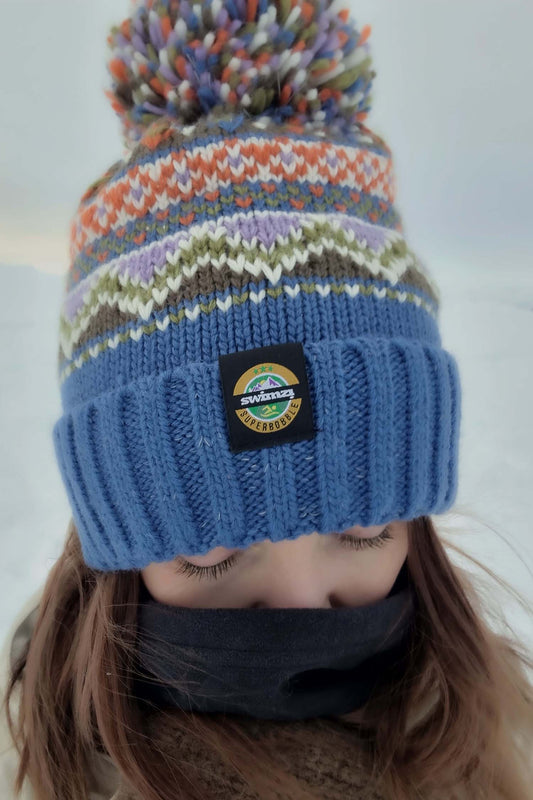 Women wearing a bobble hat with an ocean blue band and farisle knit design in  shades of olive green, lilac, orange and white.  The hat also has a deep ribbed headband and contains reflective yarns and a luxury sherpa fleece lining.