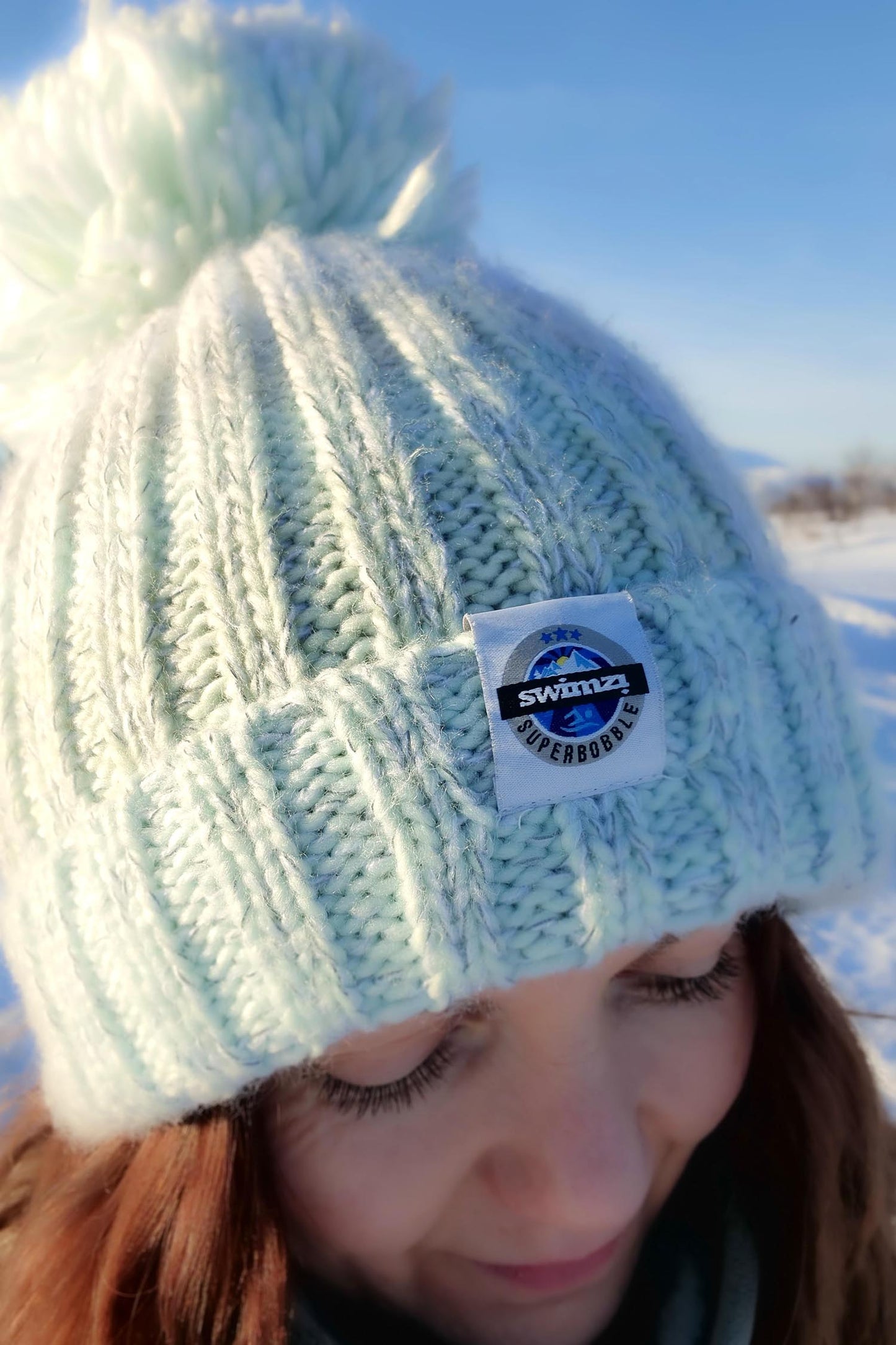  Woman standing in a snowy landscape in Norway is wearing a chunky knit Super Bobble hat in shades of peppermint . The hat has a big pom-pom and a deep ribbed headband. It contains reflective yarns and is lined in luxury sherpa fleece.