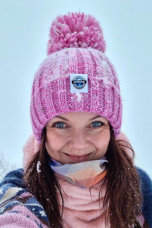 Woman standing on the edge of a snowy forest in Norway is wearing a chunky knit Super Bobble hat in shades of pink . The hat has a big pom-pom and a deep ribbed headband. It contains reflective yarns and is lined in luxury sherpa fleece.