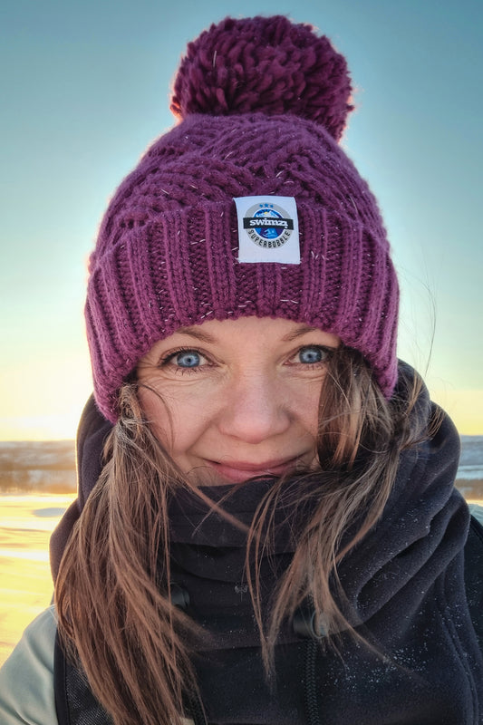 Women in a frozen landscape wearing a hat in deep rhubarb colour with a big bobble. It is knitted in chunky yarn in an attractive cross patch pattern with a deep ribbed headband. The hat contains reflective yarns and is lined in luxury sherpa fleece.