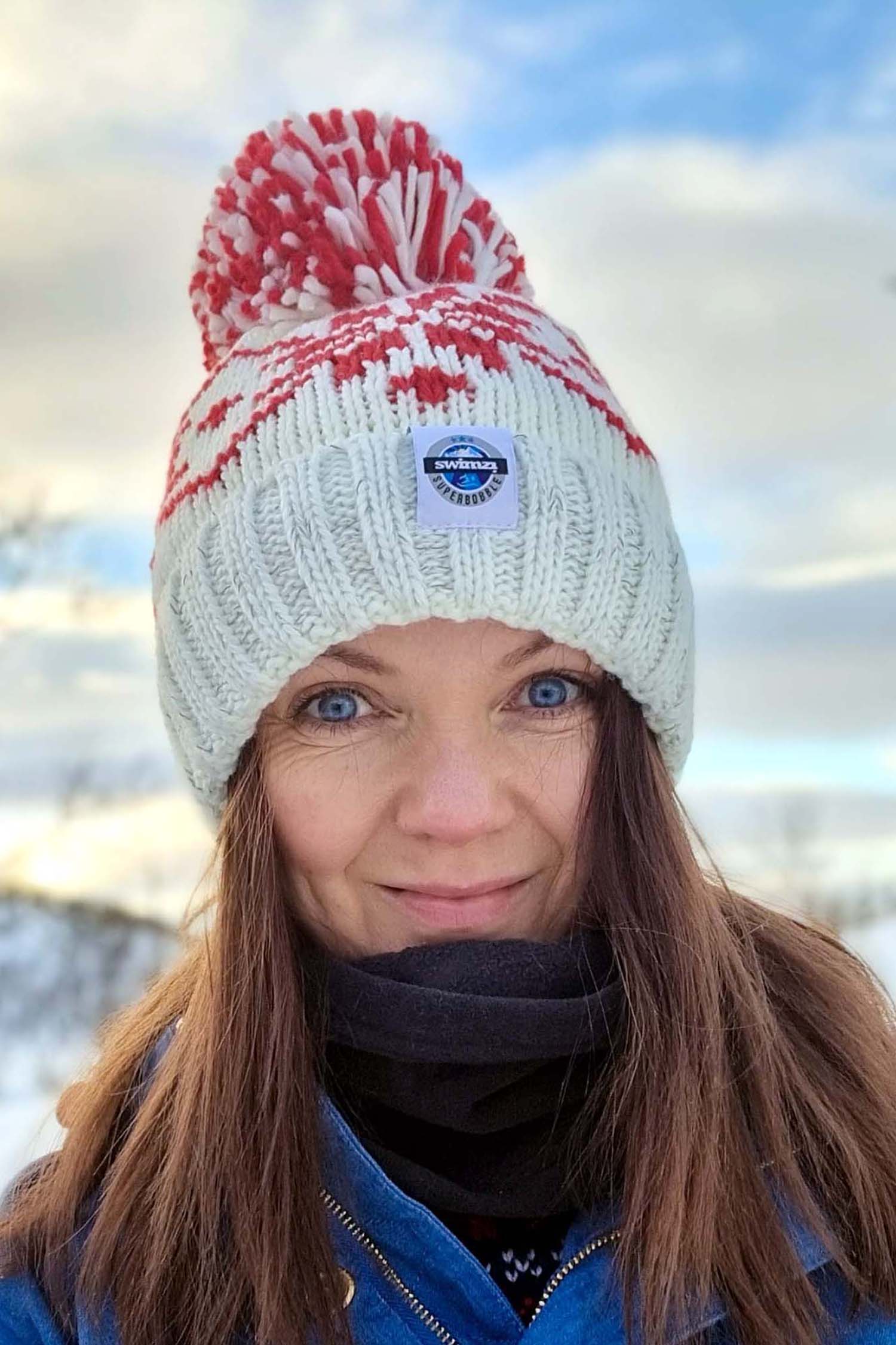 Woman in a tree lined valley is wearing a white Nordic knit bobble hat that has a repeating ruby red large snowflake around the hat and has a big pom-pom and a deep ribbed headband. It contains reflective yarns and is lined in luxury sherpa fleece.