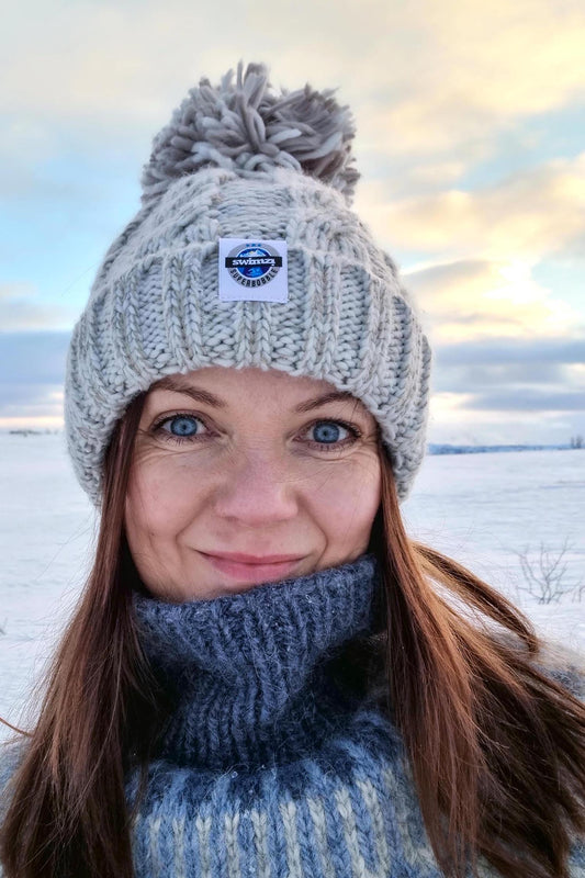 Woman standing with her dog, under a setting sun and milky blue skies is wearing a chunky cable knit silver grey Super Bobble hat. The hat has a large pom• pom, deep ribbed headband and contains reflective yarns and is lined with soft sherpa fleece.