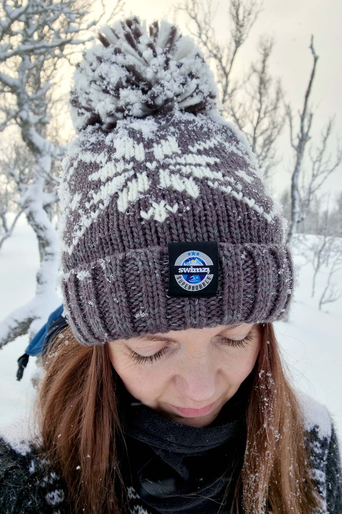  Woman on edge of snowy wood is wearing a dark grey Nordic knit bobble hat that has a repeating large white snowflake design around and has a big pom-pom and a deep ribbed headband. It contains reflective yarns and is lined in luxury sherpa fleece.