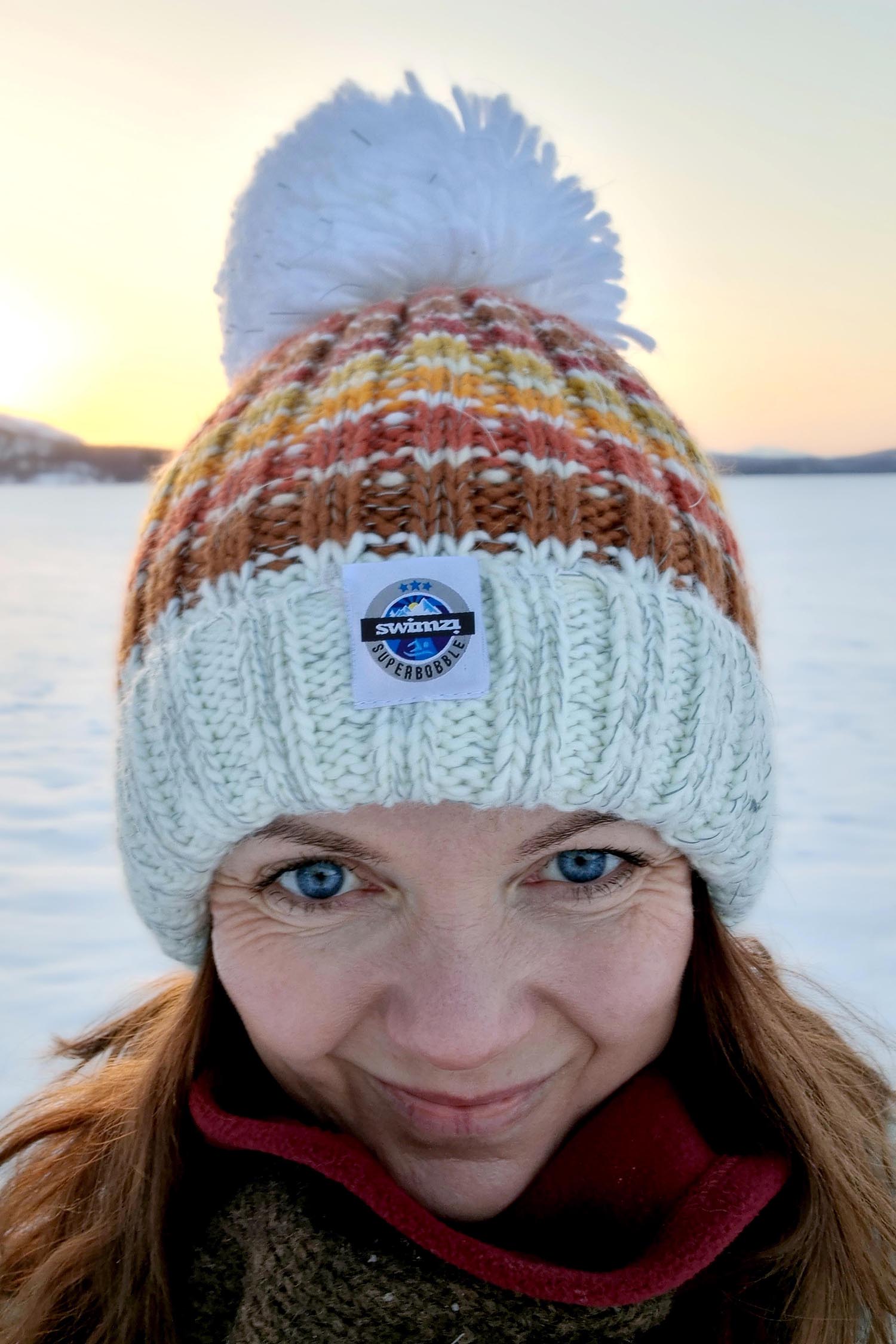 Against a snowy backdrop women wears a striped supeer bobble hat with a base colour of off white and stripes in shades of brown, burnt orange, orange and mustard. The hat also has a deep ribbed headband and a luxurious sherpa fleece lining.