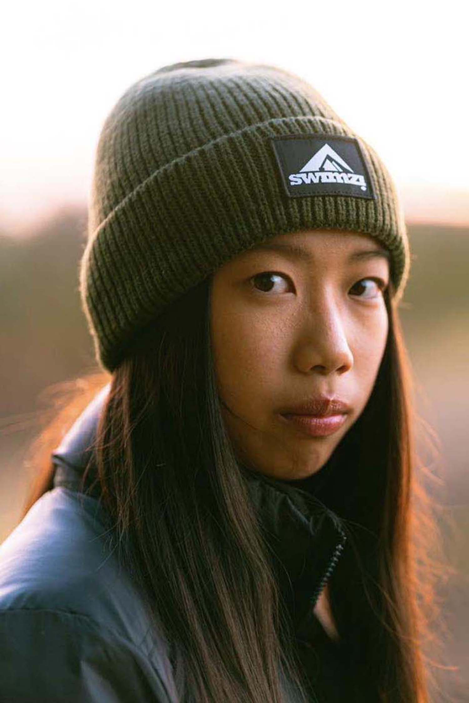 Young woman wearing a simple beanie hat knitted in a chunky single rib in a muted shade of green. There is a SWIMZI badge sewn to the front of the deep turn back headband.