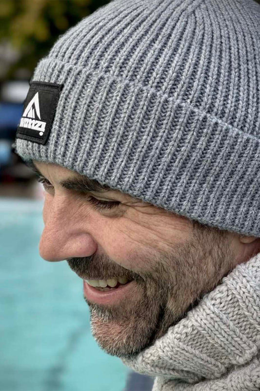 Man wearing a simple beanie hat knitted in a chunky single rib in a silver grey shade. There is a SWIMZI badge sewn to the front of the deep turn back headband.