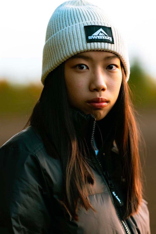 Young woman wearing a simple beanie hat knitted in a chunky single rib in an oatmeal shade. There is a SWIMZI badge sewn to the front of the deep turn back headband.