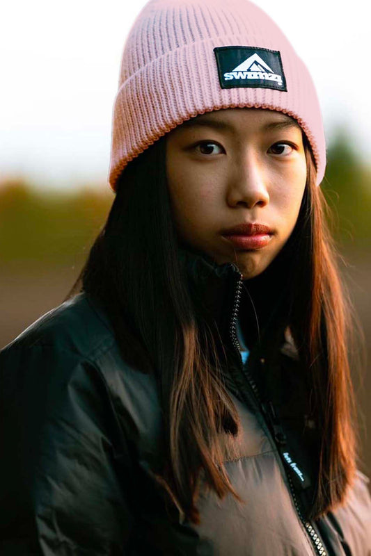 Young woman wearing a simple beanie hat knitted in a chunky single rib in a flamingo pink shade. There is a SWIMZI badge sewn to the front of the deep turn back headband.