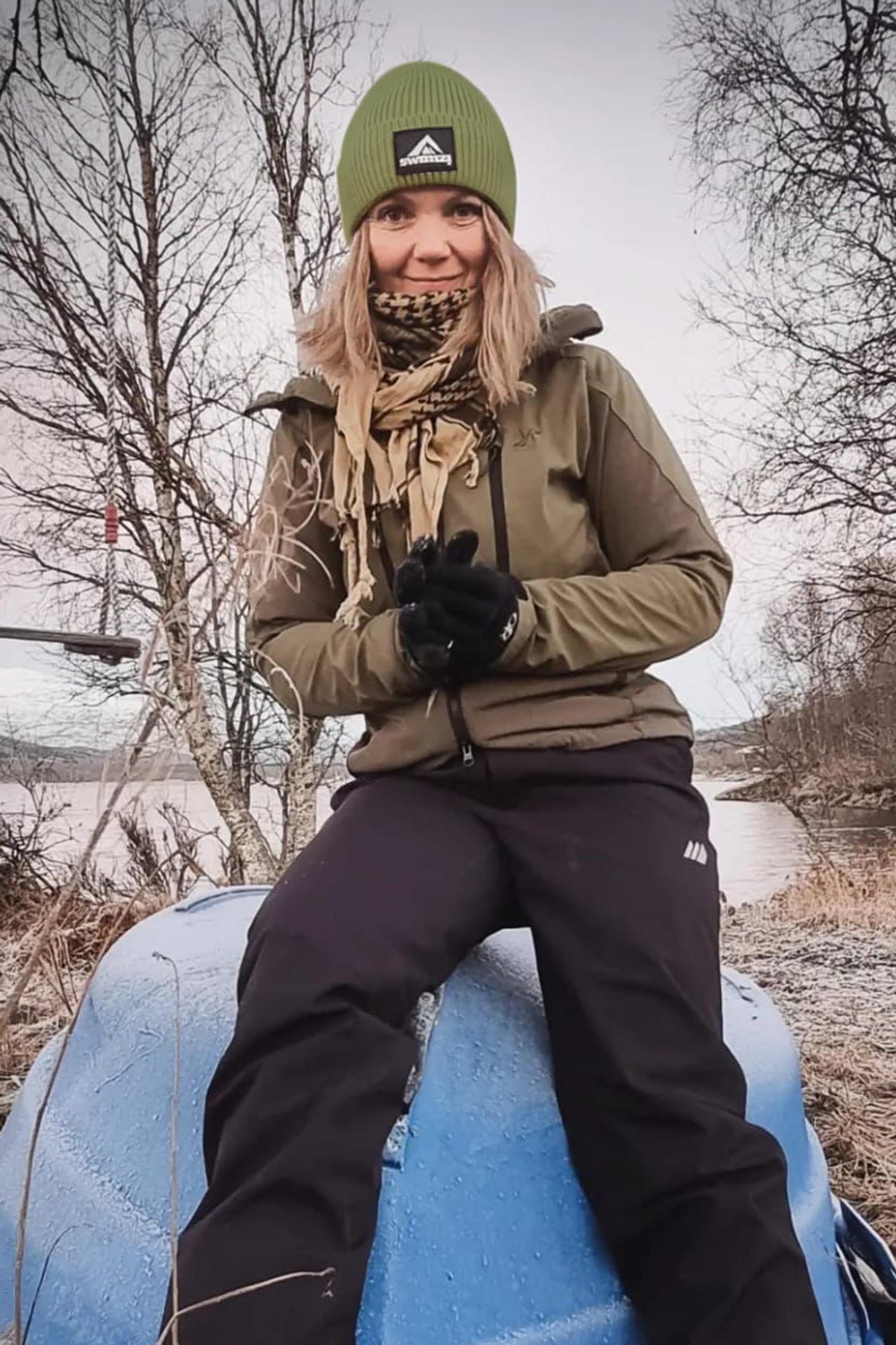 Young woman sitting on the hull of an upturned rowing boat wearing a simple beanie hat knitted in a chunky single rib in a pistachio green shade. There is a SWIMZI badge sewn to the front of the deep turn back headband.