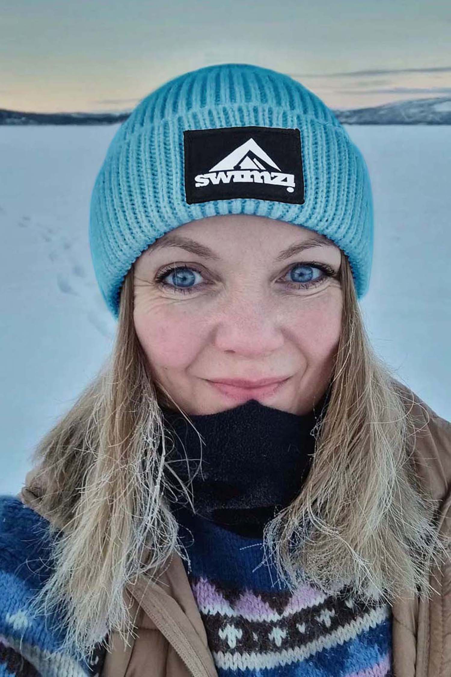 Woman wearing a simple beanie hat knitted in a chunky single rib in a sky blue shade against the snowy backdrop. There is a SWIMZI badge sewn to the front of the deep turn back headband.