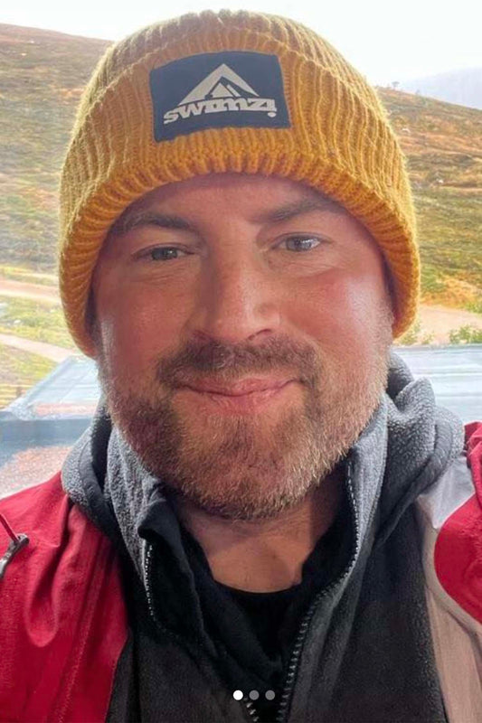 Man wearing a simple beanie hat knitted in a chunky single rib in a golden yellow shade. There is a SWIMZI badge sewn to the front of the deep turn back headband.