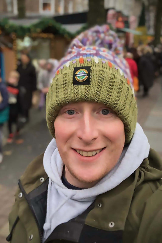 Man at a Christmas market wearing a chunky fairisle knit  bobble hat in a base of tundra green, deep ochre, aubergine and cream. The hat has a big pom-pom and a deep ribbed headband and contains reflective yarns and a luxury sherpa fleece lining.