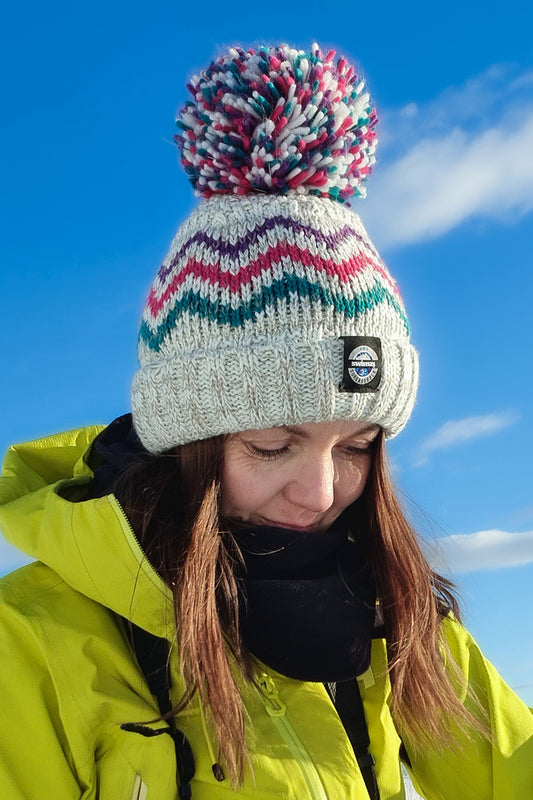 Women in a snowy landscape is wearing a chunky knit bobble hat with pink and teal peaks pattern on a white and pale grey base with a deep ribbed headband. The hat also contains reflective yarns and has a luxury sherpa fleece lining.